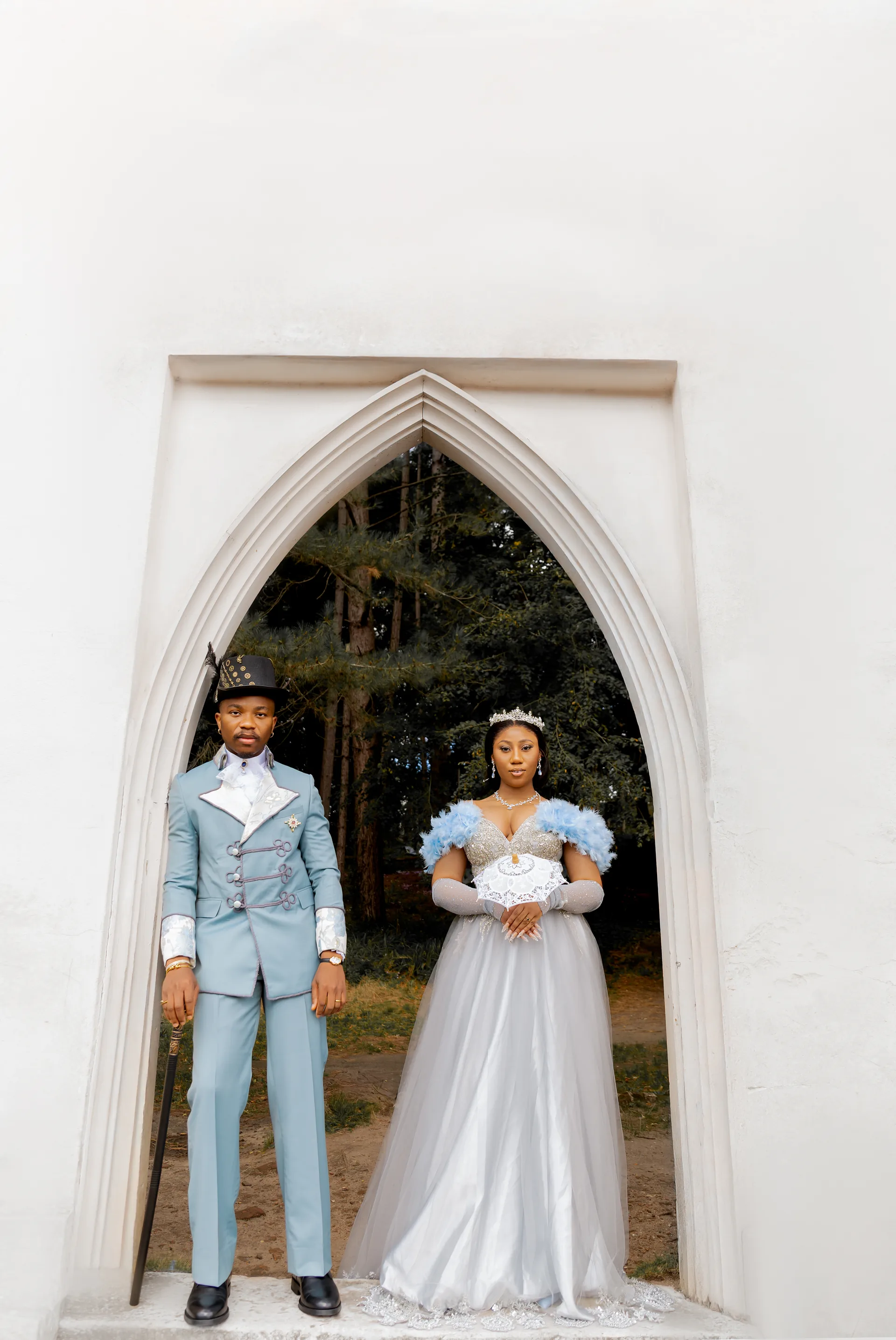 Bride And Groom In Stylized Powder Blue Outfits Posing Within A White Pointed Archway.